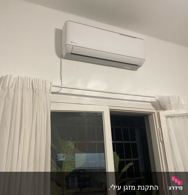 מזגן מותקן על קיר מעל חלון עם וילון לבן